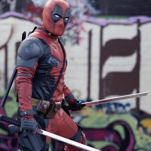 May include: A person in a red and black Deadpool costume, complete with a mask, gloves, and utility belt. They are holding two silver swords. The background features graffiti art.