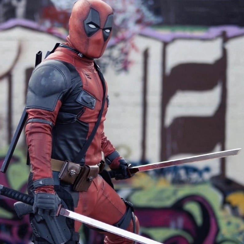 May include: A person in a red and black Deadpool costume, complete with a mask, gloves, and utility belt. They are holding two silver swords. The background features graffiti art.