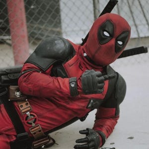 May include: A red and black Deadpool costume, featuring a mask with white eye shapes, black gloves, and shoulder pads. The character is pointing with a gloved hand, with a sword strapped to the back.