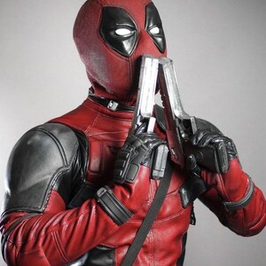 May include: A red and black Deadpool costume with a mask, holding two silver toy guns. The costume features a textured red leather-like material with black accents on the shoulders and gloves. The mask has white eye cutouts.