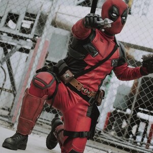 May include: A person in a Deadpool costume, kneeling and aiming two silver toy guns. The costume is red and black with a mask. The person is wearing black boots and a brown belt with pouches. The background includes a chain-link fence.