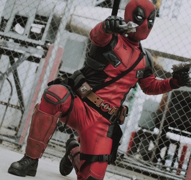 May include: A person in a Deadpool costume, kneeling and aiming two silver toy guns. The costume is red and black with a mask. The person is wearing black boots and a brown belt with pouches. The background includes a chain-link fence.