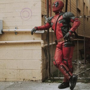 May include: A red and black Deadpool costume, including a mask, gloves, and boots. The costume features a red and black color scheme with a utility belt and a sword on the back. The person is leaning against a brick wall.