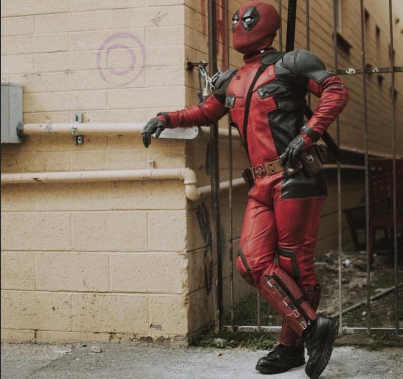 May include: A red and black Deadpool costume, including a mask, gloves, and boots. The costume features a red and black color scheme with a utility belt and a sword on the back. The person is leaning against a brick wall.