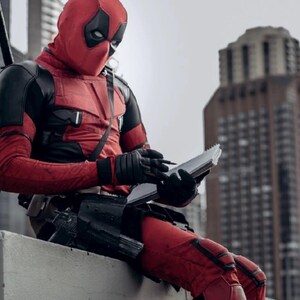 May include: A person in a red and black costume, with a mask covering the face, is writing in a notebook. The costume includes gloves, a utility belt, and a sword on the back. The background features a tall building and a cloudy sky.