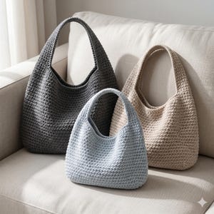 Puede incluir: Tres bolsos tote de crochet hechos a mano en varios tamaños y colores. El bolso más grande es gris, el mediano es beige claro y el más pequeño es azul claro. Todos los bolsos tienen forma redondeada y una sola correa para el hombro.