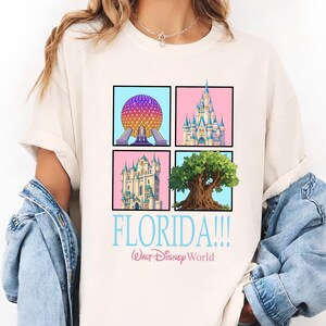 May include: Cream-colored t-shirt featuring four square illustrations of Florida landmarks. The shirt includes the text "FLORIDA!!!" in light blue, with "Walt Disney World" below in a smaller font. The illustrations include a sphere, a castle, and a tree.