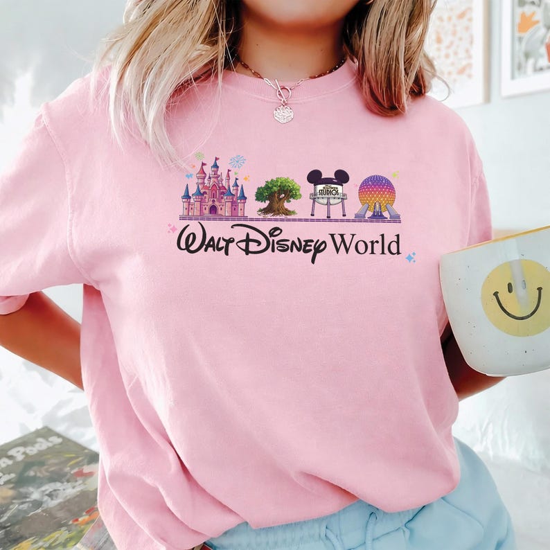 May include: Pink sweatshirt featuring a colorful graphic of Walt Disney World icons, including Cinderella's castle, a tree, Mickey Mouse ears, and Spaceship Earth. The words "Walt Disney World" are printed below the graphic in a script font. A white mug with a smiley face is visible.