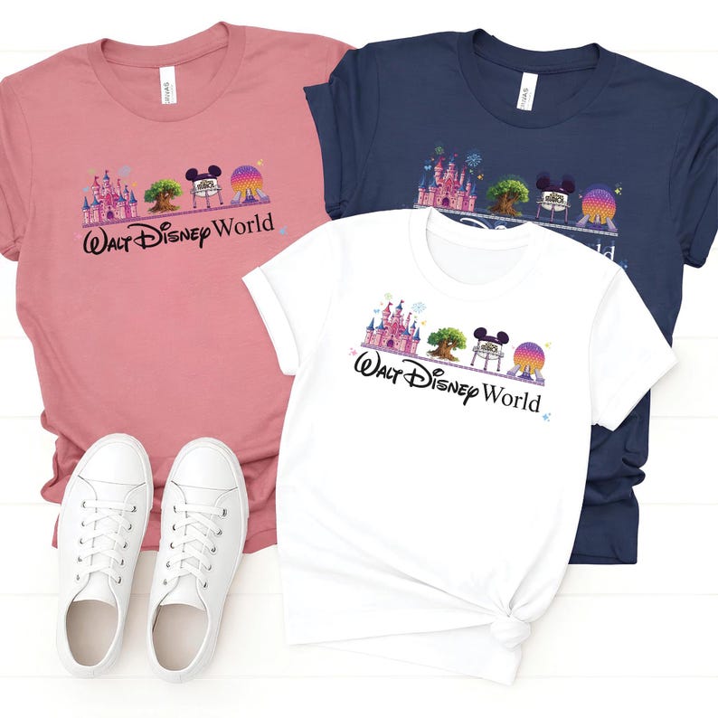 May include: Three t-shirts in pink, white, and navy blue, each featuring a colorful graphic of a castle, tree, Mickey Mouse, and a globe, with the text "Walt Disney World" in black script. White sneakers are visible.