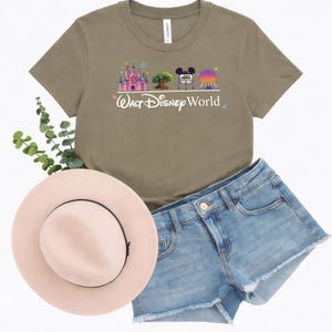May include: An olive green t-shirt with a graphic of a castle, tree, Mickey Mouse head, and a sphere, with the words "Walt Disney World" in white. The shirt is paired with denim shorts and a pink hat.