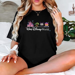 May include: Black t-shirt featuring a colorful graphic of Disney World icons, including a castle, tree, and Spaceship Earth. The words "Walt Disney World" are written in white script below the graphic. The shirt is a classic crew neck.