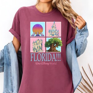 May include: Burgundy t-shirt featuring a four-square graphic with iconic Florida landmarks. The design includes a colorful sphere, a castle, a tree, and another castle, with the word "FLORIDA!!!" and "Walt Disney World" in light blue.