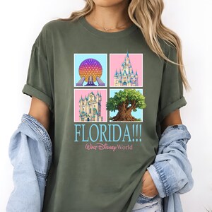 May include: Olive green t-shirt featuring four square images of Florida landmarks, including a sphere, a castle, and a tree. The word "FLORIDA!!!" is printed in blue, with "Walt Disney World" below.