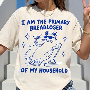 May include: Ivory-colored t-shirt with the text "I AM THE PRIMARY BREADLOSER OF MY HOUSEHOLD" in blue. The graphic features a cartoon mouse in sunglasses, holding a cocktail and a bottle, lounging on a beach chair. The shirt is a casual, unisex style.