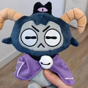 May include: A plush toy of a cartoon character with a dark blue head, tan horns, and a purple cape. The character has a grumpy expression with crossed-out eyes and a small black crown with a purple eye. The toy is held in a person's hand.