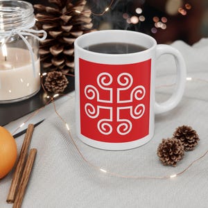 May include: White ceramic mug with a red square featuring a white, stylized cross design. The mug is filled with a dark liquid and has a white handle. The mug is on a light gray surface with decorative elements.