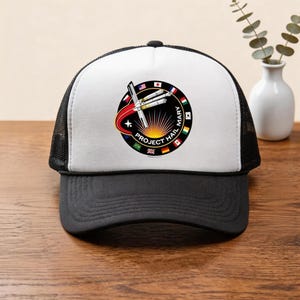 以下が含まれることがあります： 白のフロントパネルに円形のパッチが付いた、黒と白のトラッカーハット。パッチには「PROJECT HAIL MARY」の文字と旗が付いたロケットのデザインが施されています。帽子には黒いブリムとメッシュの背面が付いています。