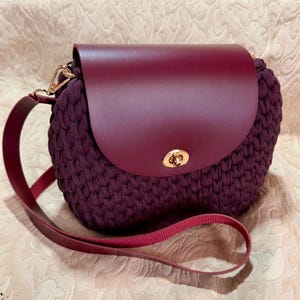 May include: A burgundy crochet crossbody bag with a flap closure. The bag features a leather flap and strap in a matching burgundy color. The bag has a gold-colored clasp and hardware. The bag is a stylish accessory.