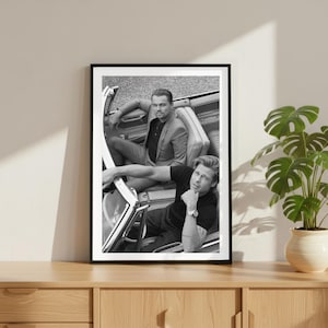 Könnte beinhalten: Schwarz-Weiß-Druck in einem Rahmen mit zwei Personen in einem Cabrio. Eine Person trägt einen Anzug, die andere ein schwarzes T-Shirt. Der Druck befindet sich auf einem Holzschrank mit einer Topfpflanze.