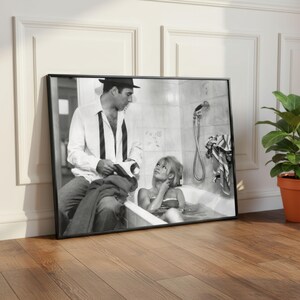 May include: Black and white framed print of a vintage photograph. The image depicts a man in a hat and a woman in a bathtub. The man is holding a book, and the woman is looking up at him. The print is leaning against a white wall.