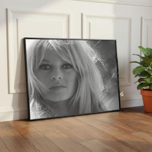 Pode incluir: Retrato emoldurado em preto e branco de uma mulher com cabelo loiro comprido. O retrato est&aacute; encostado a uma parede branca e a um ch&atilde;o de madeira. Uma planta em vaso est&aacute; &agrave; direita da moldura.