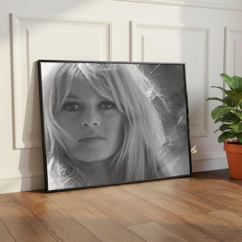 Pode incluir: Retrato emoldurado em preto e branco de uma mulher com cabelo loiro comprido. O retrato est&aacute; encostado a uma parede branca e a um ch&atilde;o de madeira. Uma planta em vaso est&aacute; &agrave; direita da moldura.