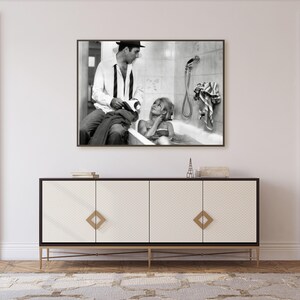 May include: Black and white framed print of a vintage photograph. The photograph depicts a man in a hat and a woman in a bathtub. The print hangs above a white cabinet with gold accents and legs. A small Eiffel Tower figurine sits on the cabinet.