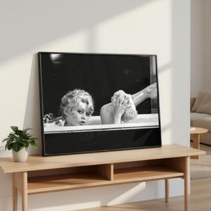 Puede incluir: Impresi&oacute;n enmarcada en blanco y negro que representa a una mujer en una ba&ntilde;era. La imagen tiene una est&eacute;tica vintage, mostrando el rostro y la parte superior del cuerpo de la mujer. Otra persona se lava la pierna con una esponja.