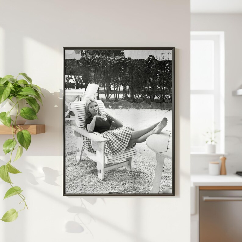 May include: Framed black and white print featuring a woman reclining in a wooden chair. She is dressed in a dark top and a checkered skirt. The scene is set outdoors, with trees in the background, creating a vintage feel.
