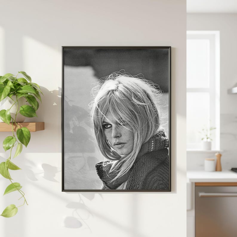 K&ouml;nnte beinhalten: Schwarz-wei&szlig; gerahmtes Portr&auml;t einer Frau mit blonden Haaren und einem Schal. Das Portr&auml;t h&auml;ngt an einer wei&szlig;en Wand neben einer Zimmerpflanze. Das Bild ist eine Nahaufnahme des Gesichts der Frau.