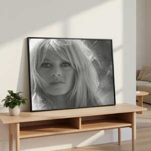 Pode incluir: Impress&atilde;o emoldurada em preto e branco de uma mulher com cabelo loiro comprido e solto. O retrato &eacute; exibido em uma mesa de console de madeira clara, com uma pequena planta em vaso &agrave; esquerda. O quarto tem uma paleta de cores neutras.