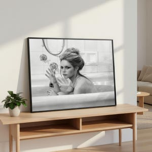 May include: Black and white framed print of a woman in a bathtub. The woman is holding a bottle and looking at the viewer. The print is displayed on a light wood console table with a small potted plant.