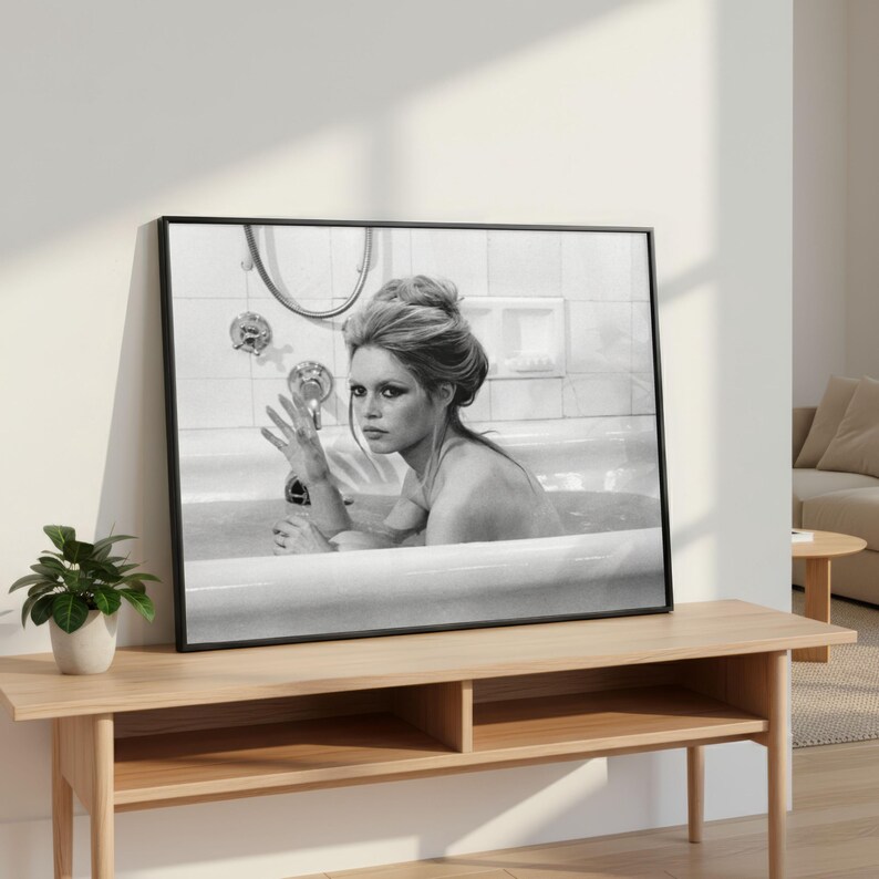 May include: Black and white framed print of a woman in a bathtub. The woman is holding a bottle and looking at the viewer. The print is displayed on a light wood console table with a small potted plant.