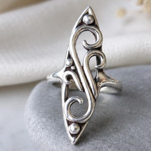 May include: A silver ring featuring an elongated, pointed oval shape with an openwork design of swirling scroll patterns. The metal surface is polished with darkened recessed areas to highlight the intricate curves and small spherical accents.