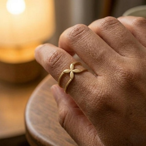 May include: A gold-colored ring with a four-petal flower design. The ring is worn on a finger, with the flower design centered on the top. The ring is made of a shiny metal and is set against a blurred background.