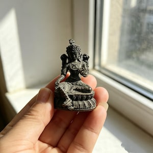 Könnte beinhalten: Eine kleine, dunkelbronze Statue einer sitzenden Gottheit. Die Figur ist mit aufwendigen Details geschmückt, darunter eine Krone, Schmuck und verzierte Kleidung. Die Statue sitzt auf einem Lotusblüten-Sockel, ein Bein ist gekreuzt. Die Statue wird in einer Hand gehalten.
