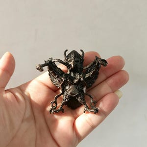 May include: A bronze-colored, detailed figurine of a mythical bird with outstretched wings, horns, and a detailed face. The bird is holding a weapon in its talons. The figurine is held in a hand.
