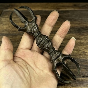 Könnte beinhalten: Ein bronzefarbener, verzierter Vajra, ein Ritualobjekt mit drei Zacken an jedem Ende, das in einer Hand gehalten wird. Der Mittelteil weist detaillierte florale und geometrische Muster auf. Der Hintergrund ist eine Holzoberfläche.