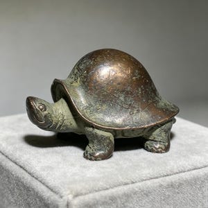 May include: A small, bronze-coloured turtle figurine with a weathered appearance. The turtle has a rounded shell, visible legs, and a smiling face. The figurine is likely made of metal and sits on a grey surface.