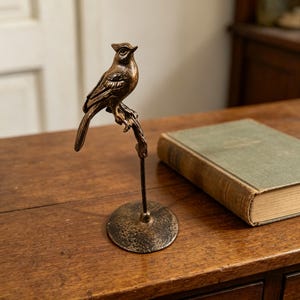 Pode incluir: Escultura de pássaro bronzeado empoleirado em um galho, montada em uma base redonda. O pássaro apresenta detalhes de penas e uma pequena crista. A escultura é exibida em uma superfície de madeira, ao lado de um livro vintage.