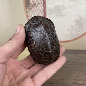 May include: A dark brown, turtle-shaped carving with intricate, engraved patterns. The detailed design covers the entire shell, showcasing a traditional aesthetic. The object is held in a hand, highlighting its size and form.