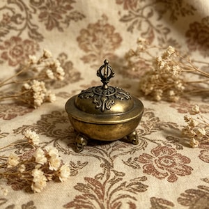 Peut inclure: Un petit brûleur d'encens en laiton antique avec un couvercle décoratif et trois pieds en forme de crâne. Le brûleur présente un design orné et une finition ternie. Le fond présente un tissu à motifs floraux.