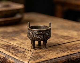 May include: A small, antique bronze incense burner with three legs and two handles. The burner features intricate, embossed patterns around the body. It sits on a weathered wooden table, showcasing its aged appearance and detailed craftsmanship.