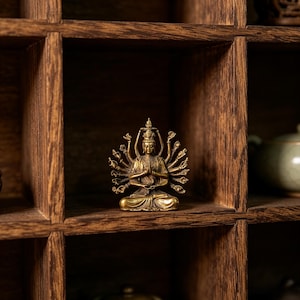 Pode incluir: Uma pequena estátua dourada de um Buda de vários braços em pose meditativa. A estátua está colocada contra um fundo de uma prateleira de madeira escura com vários compartimentos. A figura de Buda tem muitos braços e está sentada.