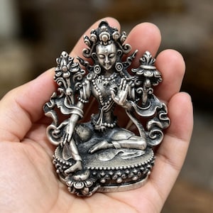 Puede incluir: Una detallada estatua plateada de una deidad sentada con elaborados tocados y joyas. La figura está en una pose meditativa, con las manos en un gesto de bendición. La estatua está sobre una base decorativa.