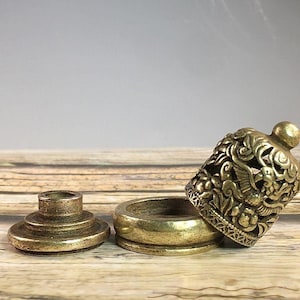 Pode incluir: Um conjunto de três peças decorativas antigas em latão. O conjunto inclui uma base, uma tampa e uma peça decorativa em forma de sino com intrincadas esculturas florais e de pássaros. As peças estão dispostas sobre uma superfície de madeira.