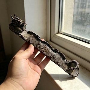 May include: A brown dragon-shaped incense holder. The holder is long and narrow, with a detailed dragon design carved along its length. The dragon's head is at one end, and its tail curls at the other. The holder is made of a dark brown material.