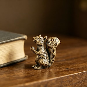 May include: A detailed brass squirrel figurine with a bushy tail and a seated pose. The squirrel is on a wooden surface, with a book visible in the background. The figurine has intricate details.