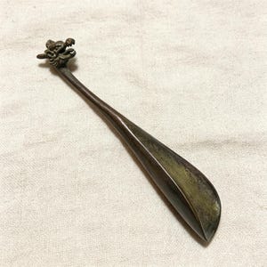 Puede incluir: Un calzador de zapatos de bronce con un diseño de cabeza de dragón en el mango. El calzador tiene un mango largo y delgado y un extremo curvado en forma de cuchara. La cabeza del dragón presenta detalles intrincados.