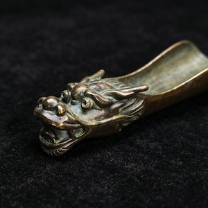 Pode incluir: Uma concha em forma de dragão, na cor bronze, com detalhes intrincados. A cabeça do dragão está na extremidade da concha, com mandíbulas abertas e escamas detalhadas. A concha tem um design curvo, em forma de calha, para servir ou medir.